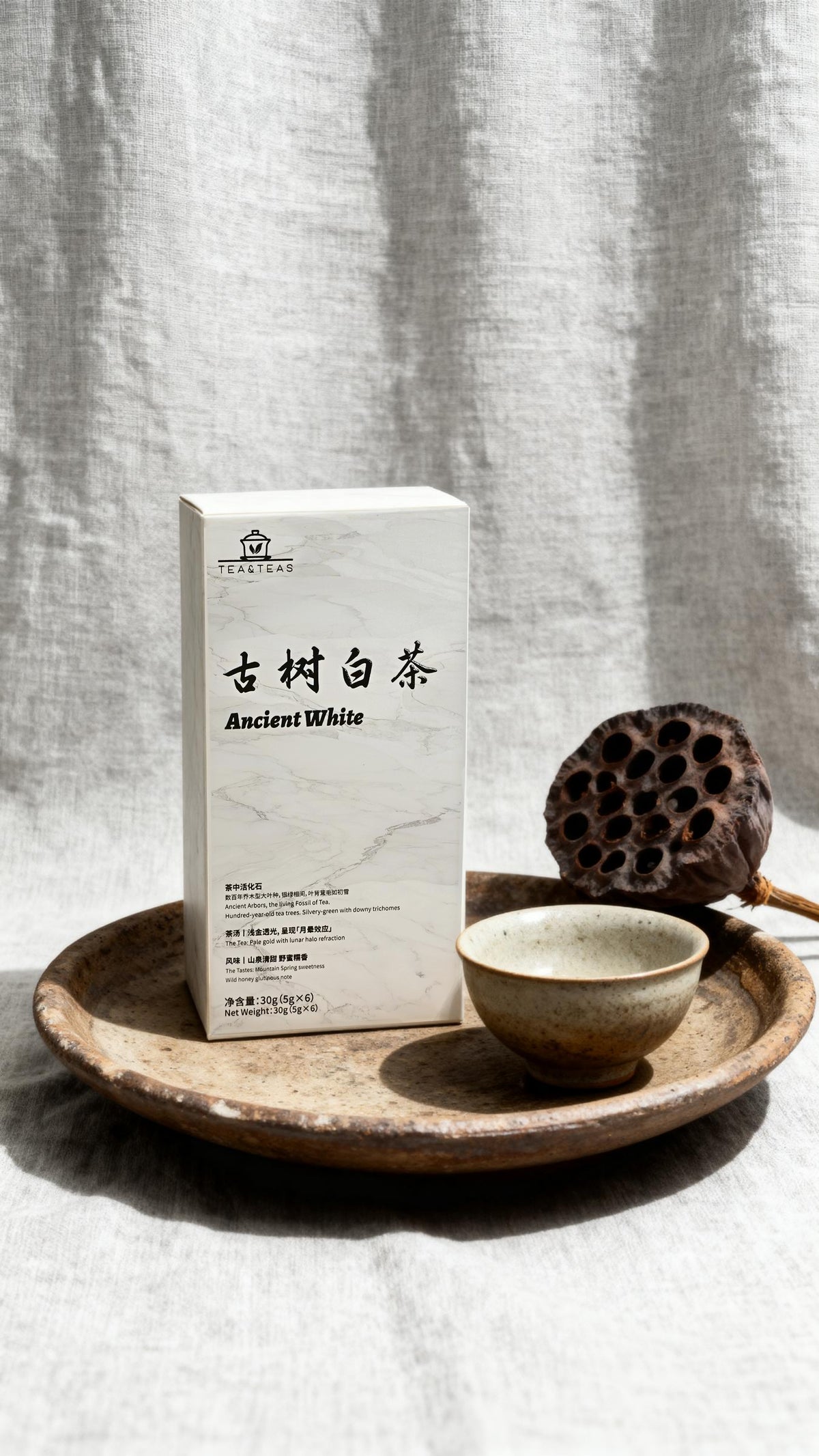 Tea box labeled 'Ancient White' on a wooden tray with a bowl and lotus seed pod against a textured gray background.